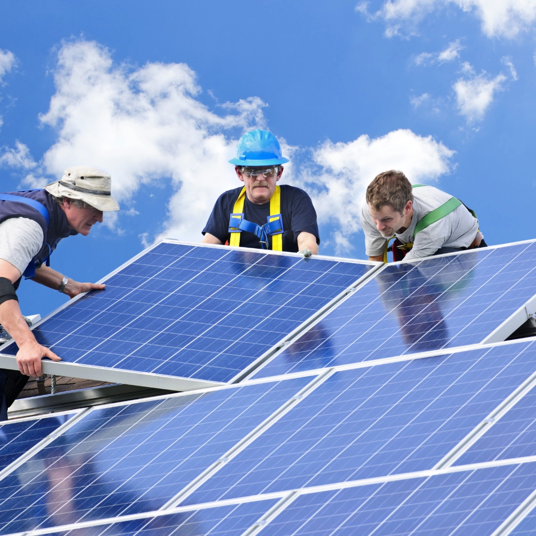 Three workers in hats and safety gear install solar panels on a roof under a bright blue sky with clouds, conveying teamwork and sustainability.